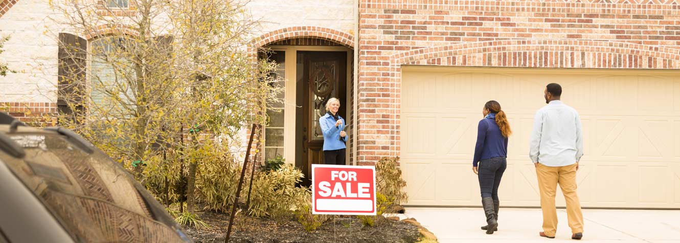 a couple approaches a realtor at a home with a for sale sign in the front yard