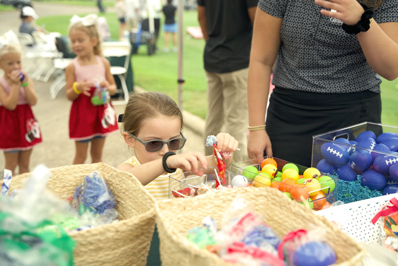 A young girl is observed by two girls in matching outfits choosing her prizes: candy, a golf ball, and a mini-football.